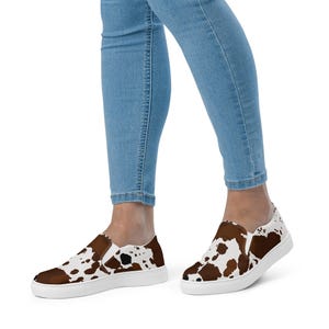 May include: Slip-on sneakers featuring a brown and white cow print pattern across the upper fabric, paired with a solid white rubber sole and white elastic side panels for a secure fit on the foot.