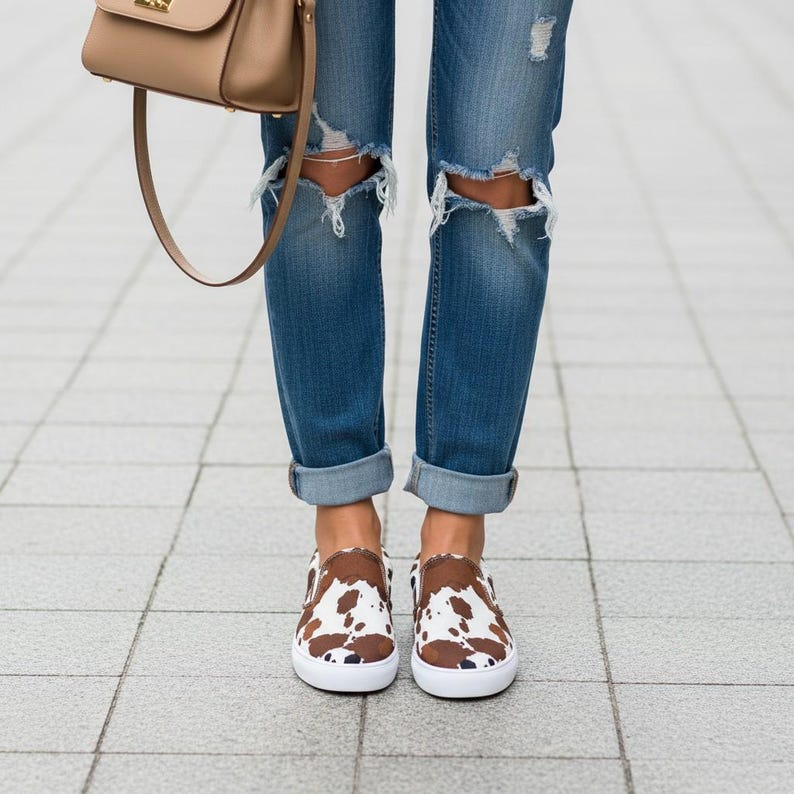 May include: A pair of slip-on sneakers featuring a brown and white cow print pattern with white rubber soles, worn with blue denim jeans that have distressed knee detailing and a tan shoulder bag with a gold-tone clasp hanging at the side.