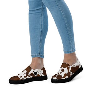 May include: Slip-on sneakers featuring a brown and white cow print pattern across the upper fabric, paired with a solid black rubber sole. The shoes are worn with light blue denim jeans, showcasing a low-top design with elastic side panels.