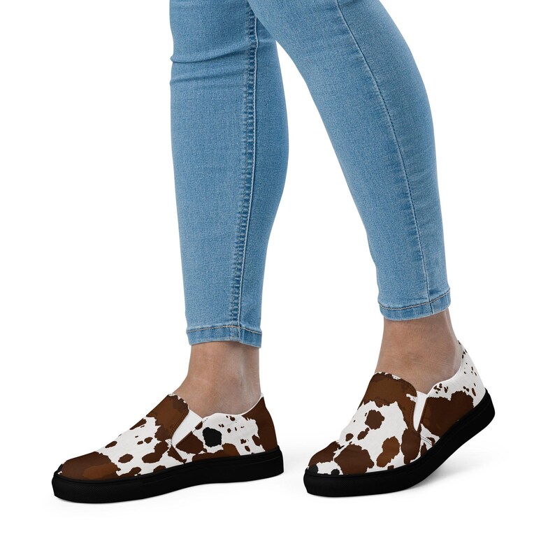 May include: Slip-on sneakers featuring a brown and white cow print pattern across the upper fabric, paired with a solid black rubber sole. The shoes are worn with light blue denim jeans, showcasing a low-top design with elastic side panels.