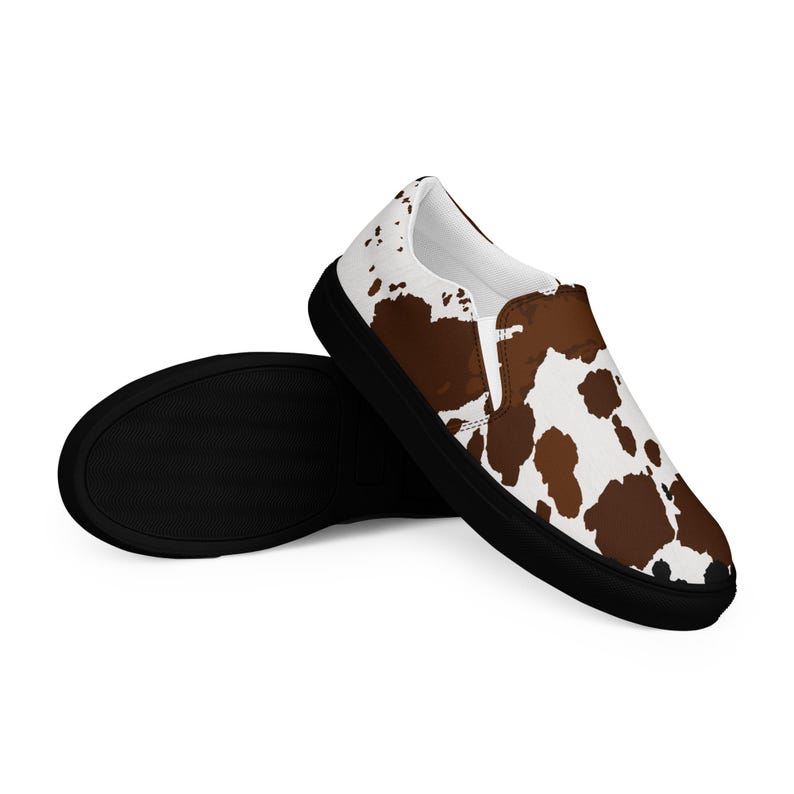 May include: A pair of slip-on sneakers featuring a white and brown cow print pattern on the upper fabric. The shoes have a solid black rubber sole and a white interior lining. The design covers the entire exterior of the shoe upper.
