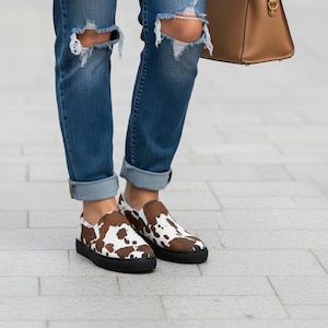 May include: Slip-on sneakers featuring a brown and white cow print pattern with solid black rubber soles, paired with blue denim jeans that have distressed knee detailing and a tan leather handbag held at the side of the legs.