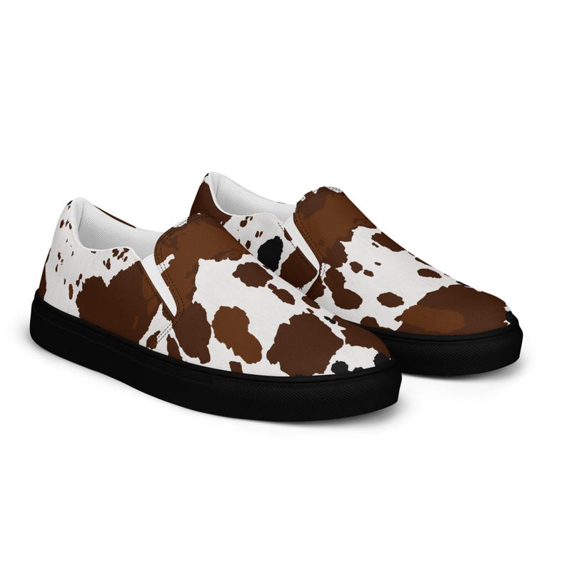 May include: A pair of slip-on sneakers featuring a brown and white cow print pattern across the upper fabric. The shoes have a solid black rubber sole and a white interior lining, designed as a low-top style with no laces or visible fasteners.