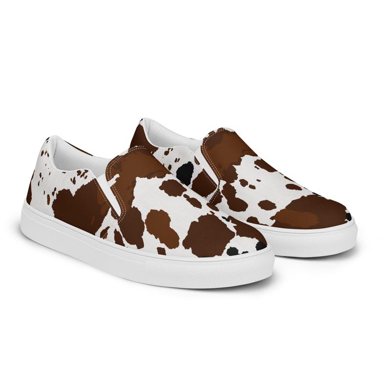 May include: A pair of slip-on sneakers featuring a brown, white, and black cow print pattern across the canvas upper. The shoes have a white rubber sole, a rounded toe, and elastic side panels for a secure fit without laces or fasteners.