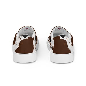 May include: A pair of slip-on sneakers featuring a brown and white cow print pattern across the exterior fabric. The shoes have a solid white rubber sole and a white interior lining with a visible brand label on the heel area of the insole.