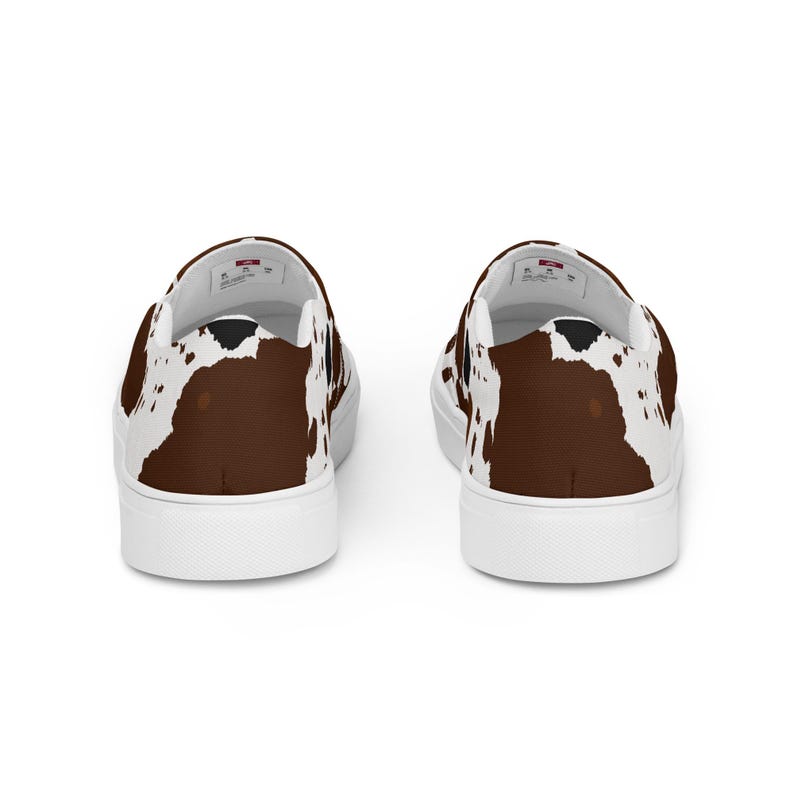 May include: A pair of slip-on sneakers featuring a brown and white cow print pattern across the exterior fabric. The shoes have a solid white rubber sole and a white interior lining with a visible brand label on the heel area of the insole.