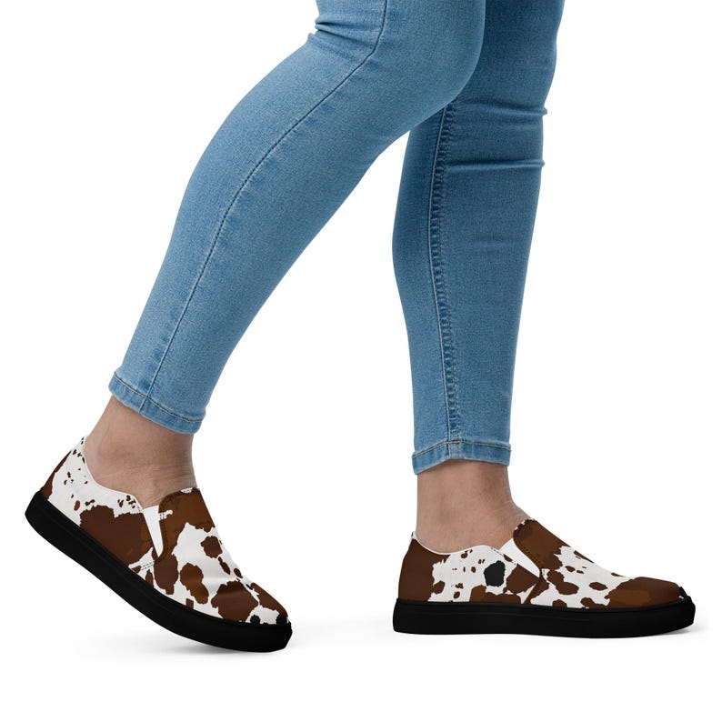 May include: A pair of slip-on shoes featuring a brown and white cow print pattern across the upper fabric. The shoes have a solid black rubber sole and a low-profile design, shown on feet wearing light blue denim jeans.