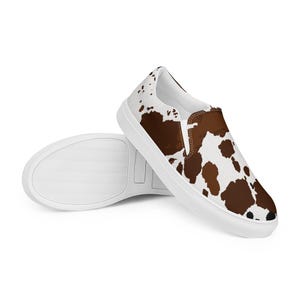 May include: A pair of slip-on shoes featuring a white base with irregular brown cow-print spots. The shoes have a low-top design with a white rubber sole and a white interior lining, showcasing a clean, laceless construction for easy wear.