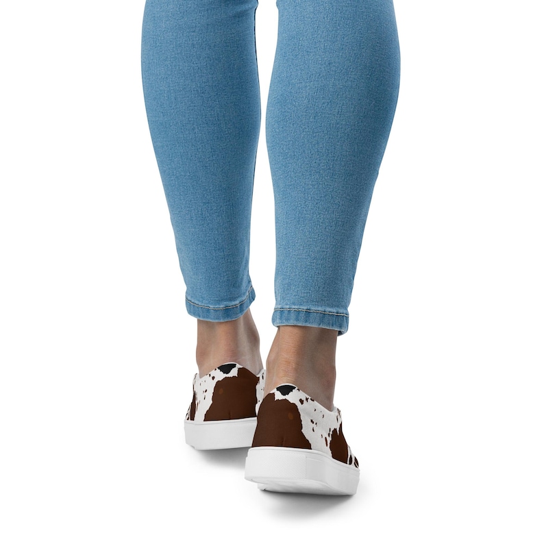 May include: Low-top sneakers featuring a brown and white cow print pattern across the upper. The footwear has a solid white rubber sole and a rounded toe design. The shoes are worn with light blue denim jeans that reach down to the ankles.