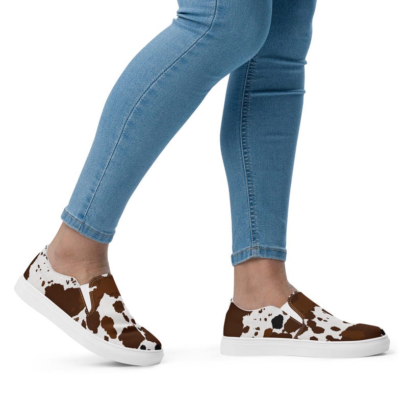 May include: A pair of slip-on sneakers featuring a brown and white cow print pattern across the upper fabric. The shoes have a low-top silhouette with white rubber soles and elastic side panels, worn with light blue denim jeans.