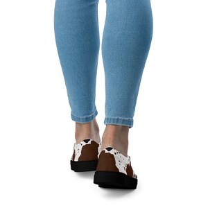 May include: A pair of slip-on shoes featuring a brown and white cow print pattern with solid black soles, worn with light blue denim jeans. The shoes have a low-top design and a rounded toe, showcasing a distinct spotted animal hide aesthetic.