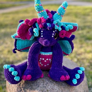 Könnte beinhalten: Ein lila gehäkelter Drache mit türkisfarbenen und rosafarbenen Akzenten. Der Drache hat große, funkelnde Augen und sitzt auf einem Stein.
