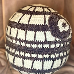 May include: A large, round, crocheted pouf in cream and dark gray. The pouf has a striped pattern with dark gray vertical stripes and cream-colored horizontal stripes. A smaller, matching round cushion is attached to the side.