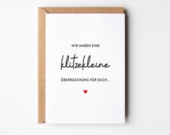 Pode incluir: Um cartão de felicitações branco com um envelope castanho apresentando o texto em alemão WIR HABEN EINE klitzekleine ÜBERRASCHUNG FÜR EUCH... em letras pretas, centrado com um pequeno ícone de coração vermelho na base.