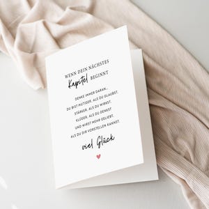 May include: A white greeting card with black text and a small pink heart. The card reads "WENN DEIN N&Auml;CHSTES Kapitel BEGINNT" and other inspirational text in German. The card is open, revealing the message, and rests on a beige fabric.