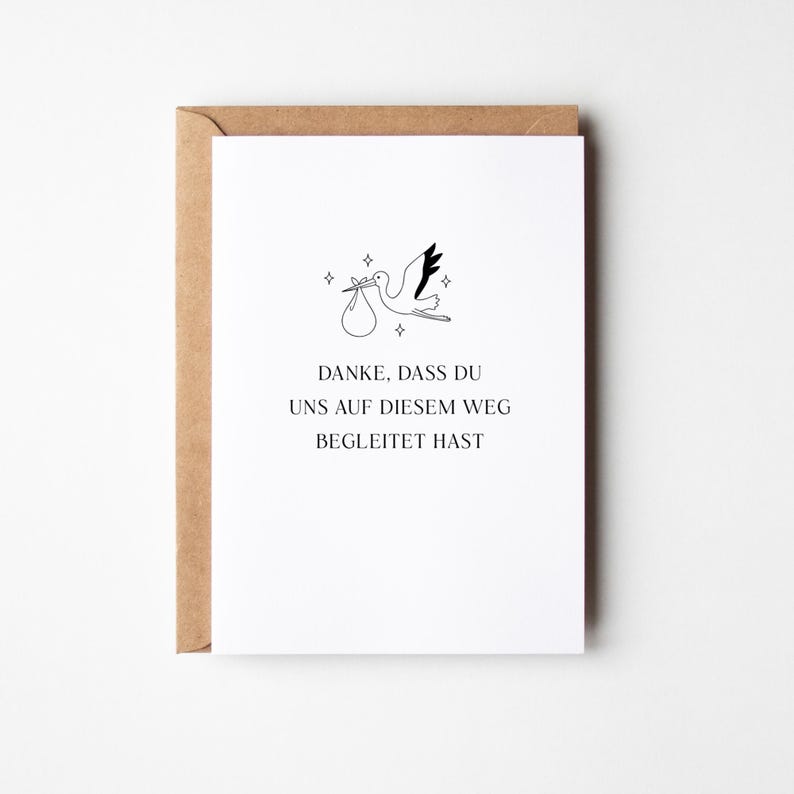 Puede incluir: Tarjeta blanca con ilustraci&oacute;n de una cig&uuml;e&ntilde;a llevando un paquete, con el texto en alem&aacute;n "DANKE, DASS DU UNS AUF DIESEM WEG BEGLEITET HAST." La tarjeta est&aacute; dentro de un sobre marr&oacute;n, ideal para un anuncio de nacimiento.