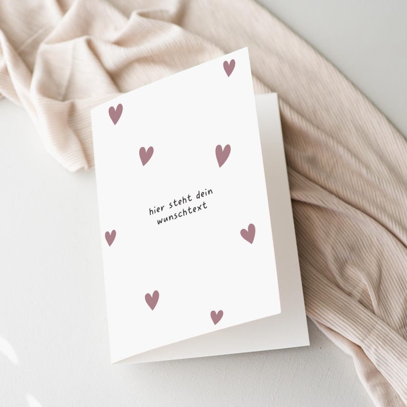 May include: A white greeting card with a design of small, mauve hearts scattered across the front. The card has the German text "hier steht dein wunschtext" in a simple font. The card is open, revealing a blank interior.