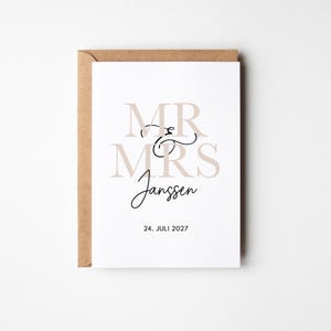 Könnte beinhalten: Eine weiße rechteckige Grußkarte mit einem braunen Kraftpapierumschlag. Die Karte zeigt den Text MR & MRS in großen beigen Serifenschriftzeichen, den Namen Janssen in schwarzer Schreibschrift und das Datum 24. JULI 2027 in schwarzer serifenloser Schrift.