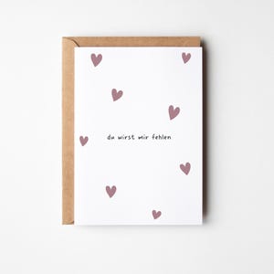 May include: A white greeting card with scattered mauve hearts and the German text "du wirst mir fehlen". The card is inside a brown envelope. The design is simple and elegant.