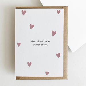 May include: A greeting card with a white background and scattered pink hearts. The card has the text "hier steht dein wunschtext" in a black font. The card is inside a brown envelope.