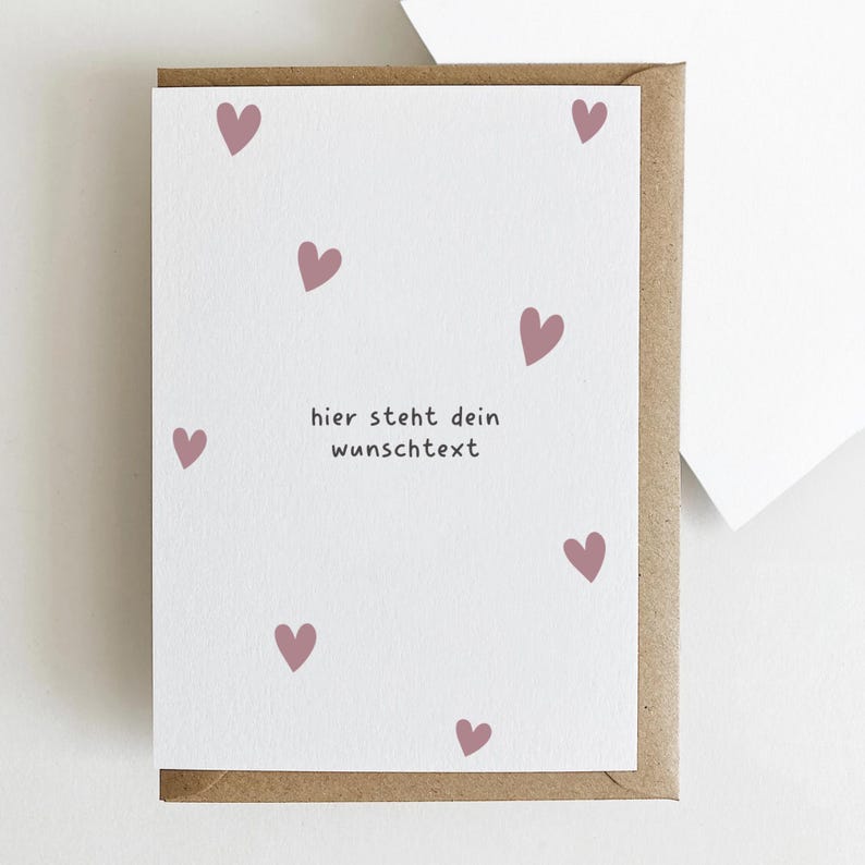 May include: A greeting card with a white background and scattered pink hearts. The card has the text "hier steht dein wunschtext" in a black font. The card is inside a brown envelope.