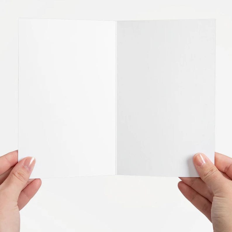 Puede incluir: Una tarjeta de felicitaci&oacute;n blanca y en blanco, abierta para mostrar el interior. La tarjeta est&aacute; sujeta por dos manos sobre un fondo blanco. La tarjeta es de tama&ntilde;o est&aacute;ndar, con un acabado liso y mate.
