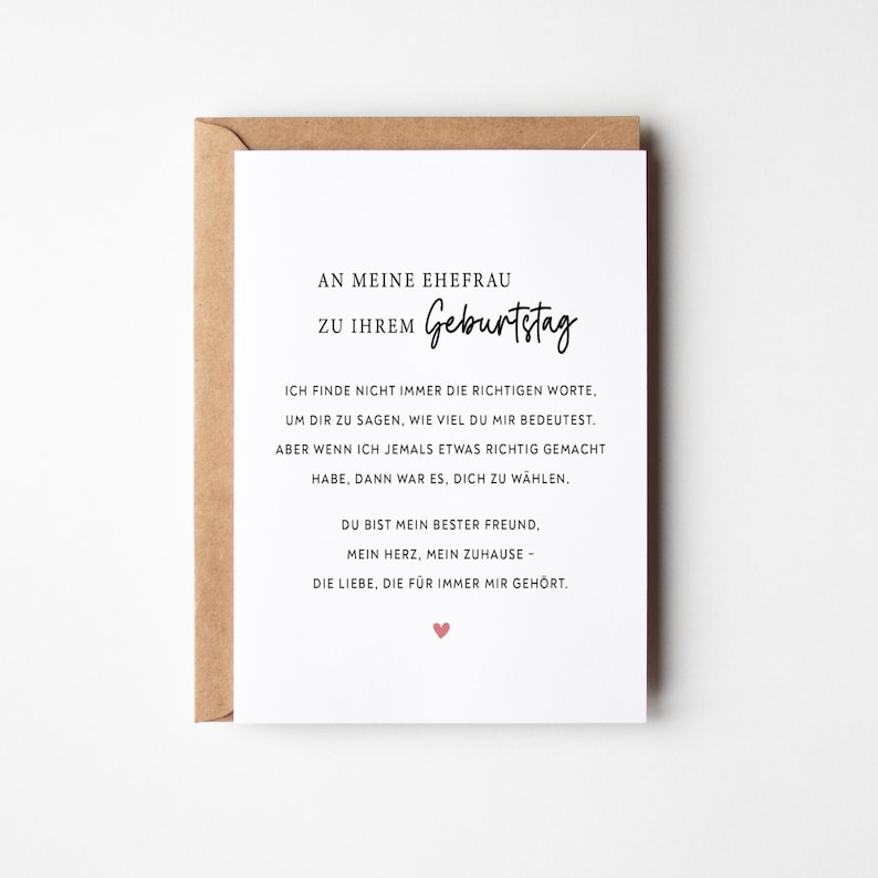 May include: A white greeting card with German text, presented in a brown envelope. The card's message is a birthday wish to a wife, with a small pink heart at the bottom. The card's text is in black, with the word "Geburtstag" in a handwritten style.