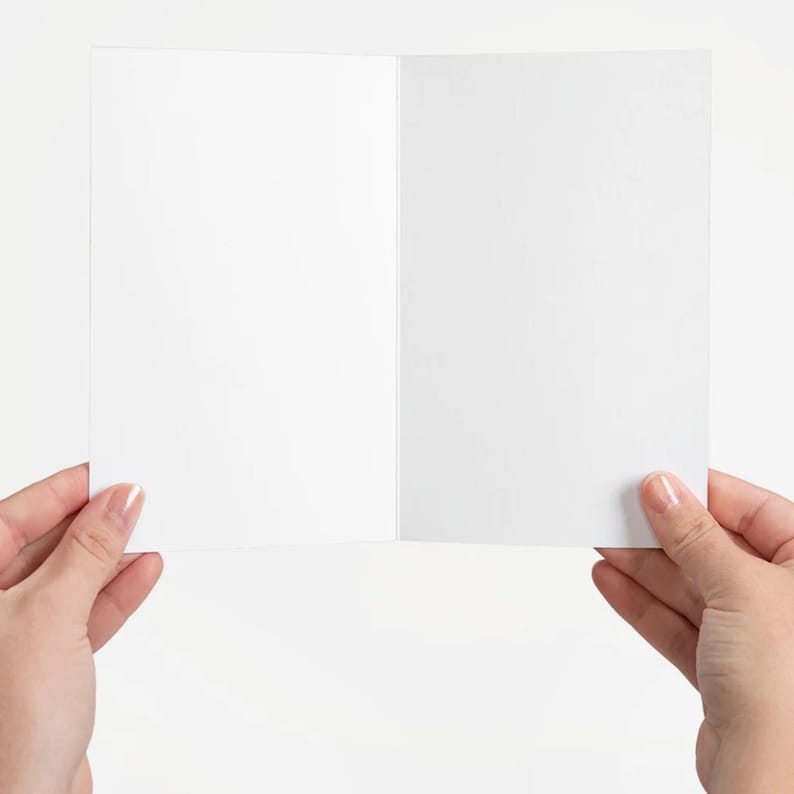 Puede incluir: Una tarjeta de felicitaci&oacute;n blanca y en blanco, abierta y sostenida por dos manos. La tarjeta est&aacute; desplegada, revelando un interior limpio y vac&iacute;o. El fondo es una superficie blanca lisa, que proporciona un tel&oacute;n de fondo neutro para la tarjeta.