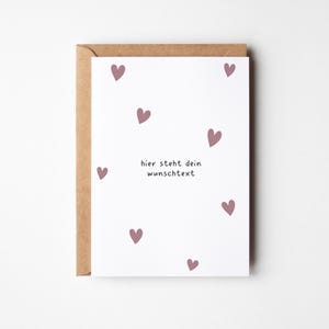 May include: A greeting card with a white background and scattered mauve hearts. The card reads "hier steht dein wunschtext" in black text. The card is inside a brown envelope.