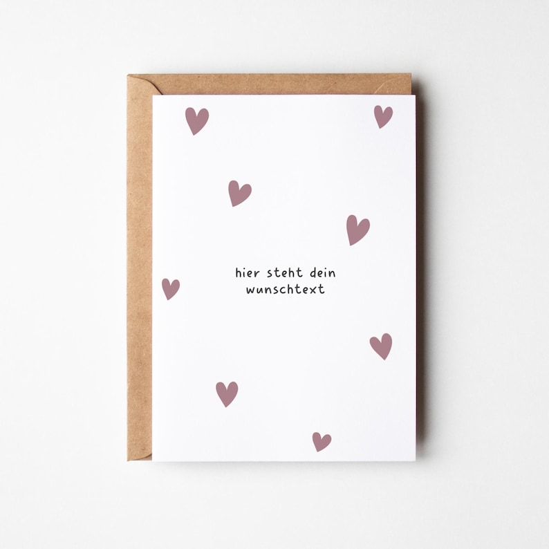 May include: A greeting card with a white background and scattered mauve hearts. The card reads "hier steht dein wunschtext" in black text. The card is inside a brown envelope.