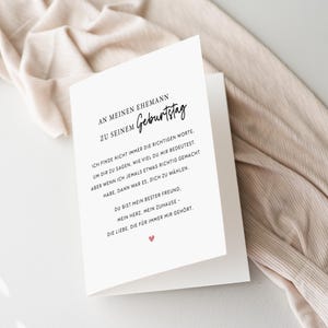 May include: A white greeting card with black text and a small red heart. The card is open, revealing a message in German. The card is resting on a beige fabric.