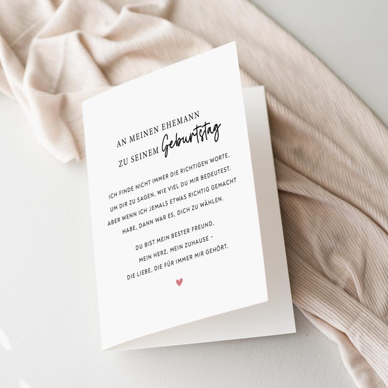 May include: A white greeting card with black text and a small red heart. The card is open, revealing a message in German. The card is resting on a beige fabric.