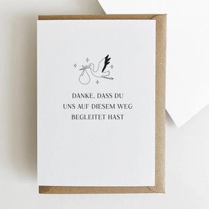 Puede incluir: Tarjeta blanca con una ilustraci&oacute;n de cig&uuml;e&ntilde;a llevando un bulto, rodeada de estrellas. La tarjeta est&aacute; en un sobre marr&oacute;n. El texto de la tarjeta dice: "DANKE, DASS DU UNS AUF DIESEM WEG BEGLEITET HAST."