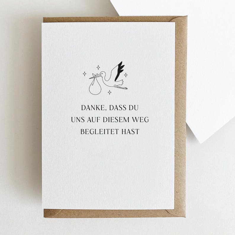 Puede incluir: Tarjeta blanca con una ilustraci&oacute;n de cig&uuml;e&ntilde;a llevando un bulto, rodeada de estrellas. La tarjeta est&aacute; en un sobre marr&oacute;n. El texto de la tarjeta dice: "DANKE, DASS DU UNS AUF DIESEM WEG BEGLEITET HAST."