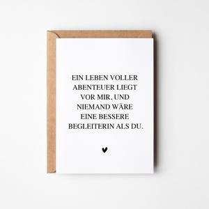 Könnte beinhalten: Eine weiße Grußkarte mit dem Text "EIN LEBEN VOLLER ABENTEUER LIEGT VOR MIR, UND NIEMAND WÄRE EINE BESSERE BEGLEITERIN ALS DU." in schwarzer Schrift und einem kleinen schwarzen Herz. Die Karte befindet sich in einem braunen Umschlag.