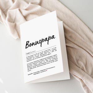 May include: A white greeting card with the word "Bonuspapa" in black script. The card is open, revealing text in German. The card is on a neutral-toned fabric background.