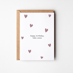 May include: A white greeting card with a brown envelope. The card features scattered mauve hearts and the text "happy birthday, liebe anna" in a simple font. The card is a standard size, perfect for sending birthday wishes.