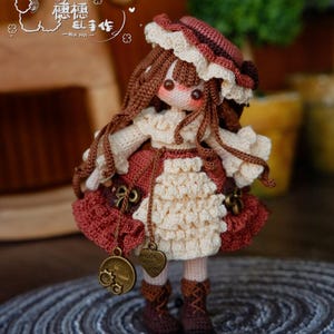 May include: A handmade crochet doll with long brown hair, wearing a red and cream-colored dress and hat. The doll has a detailed face with rosy cheeks and is adorned with small metal charms. The doll is standing on a gray, circular rug.