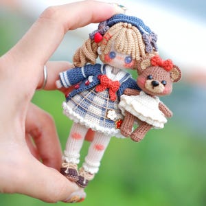 Könnte beinhalten: Eine handgefertigte Häkelpuppe mit blauem Matrosen-Outfit, kariertem Rock und roter Schleife. Die Puppe hält einen kleinen gehäkelten Teddybären. Die Puppe hat blondes Haar und braune Schuhe. Die Puppe ist etwa 13 cm groß.