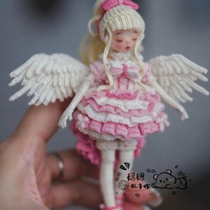 May include: A handcrafted crochet doll featuring blonde hair, white feathered wings, and a layered pink and white dress with ruffled details. The doll has closed eyes and is wearing pink lace-up shoes. A small logo with a bear graphic is visible.