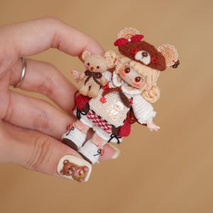 以下が含まれることがあります： 手作りのクマをテーマにした衣装と帽子を着た人形が手に持たれています。人形には小さなクマの仲間がいます。人形の衣装には、赤と白のベスト、白いブーツ、赤いリボンが付いた茶色の帽子が含まれています。