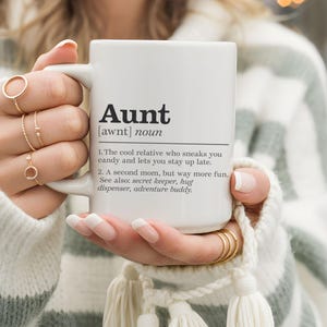 Könnte beinhalten: Weiße Keramik-Tasse mit der Definition von "Aunt" in schwarzer Schrift. Die Definition enthält Sätze wie "coole Verwandte" und "zweite Mutter". Die Tasse wird von einer Person gehalten, die goldene Ringe trägt.