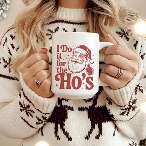 Op de afbeelding: Witte keramische mok met een rode Santa Claus-afbeelding en de tekst "I Do it for the Ho's". De mok wordt vastgehouden door iemand die een witte trui met een rendierenpatroon draagt. Een feestelijk kerstcadeau.