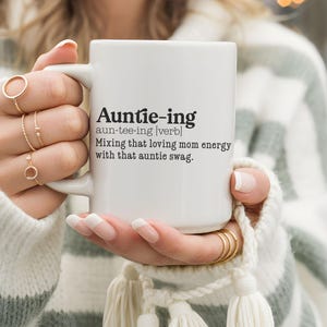 May include: White ceramic mug with the text "Auntie-ing" and its definition. The definition reads "Mixing that loving mom energy with that auntie swag." The mug is held by a person wearing gold rings.
