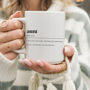May include: White ceramic mug with the definition of "aunt" printed in black. The definition includes the phrase "Like a mom, only cooler, with better gifts and no rules." The mug is held by a person wearing gold rings.