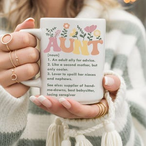 Könnte beinhalten: Weißer Keramikbecher mit dem Wort "AUNT" in bunten Buchstaben und einem floralen Design. Der Becher enthält eine Definition von "AUNT" mit drei Punkten und verwandten Begriffen. Ein perfektes Geschenk für eine Tante.