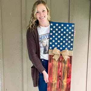 May include: A wooden American flag with a blue star field and red and brown stripes. The flag is decorated with a burlap bow. The flag is approximately 12 inches wide and 24 inches tall.