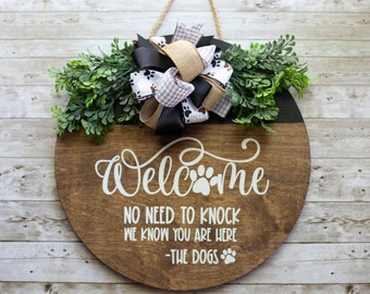 May include: A round wooden door hanger with a black and brown paint design. It says "Welcome" with a paw print in the middle. The text below says "No need to knock we know you are here -The Dogs" with a paw print at the end. A bow with black, white, and brown ribbon is attached to the top.