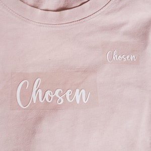 Könnte beinhalten: Ein hellrosa T-Shirt mit dem Wort "Chosen" in weiß auf der Vorderseite.