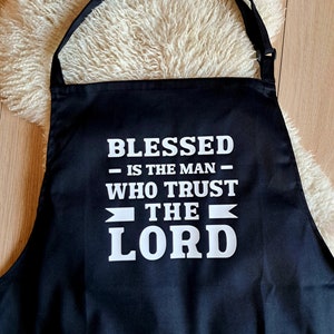 Könnte beinhalten: Schwarze Küchenschürze mit weißem Text, der "Blessed is the man who trust the Lord" lautet.